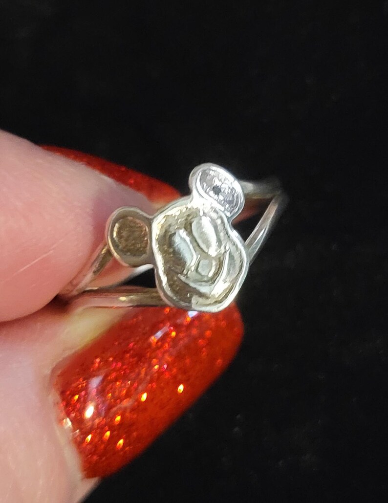 Puede incluir: Un anillo de tono plateado que presenta una cabeza de personaje estilizada en relieve con dos orejas redondeadas. El anillo tiene un dise&ntilde;o de banda dividida que se conecta al motivo central. La superficie met&aacute;lica tiene un acabado pulido.