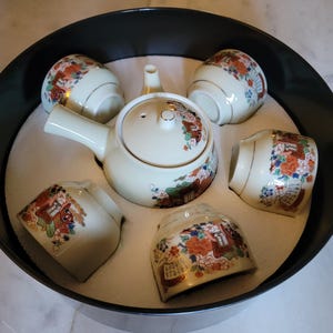 May include: A Japanese tea set with a teapot, cups, and a small vase, arranged in a black tray filled with sand. The tea set is cream-colored with floral and gold accents.