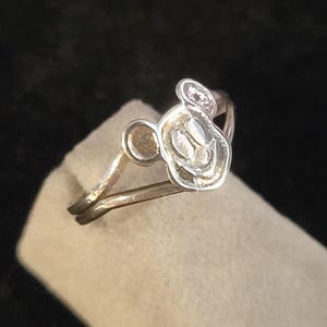 Puede incluir: Un anillo de metal plateado que presenta una cabeza de personaje estilizada en relieve con dos orejas redondeadas. La banda del anillo es delgada y se divide en dos secciones al conectar con el dise&ntilde;o central, creando un aspecto simple y abierto.