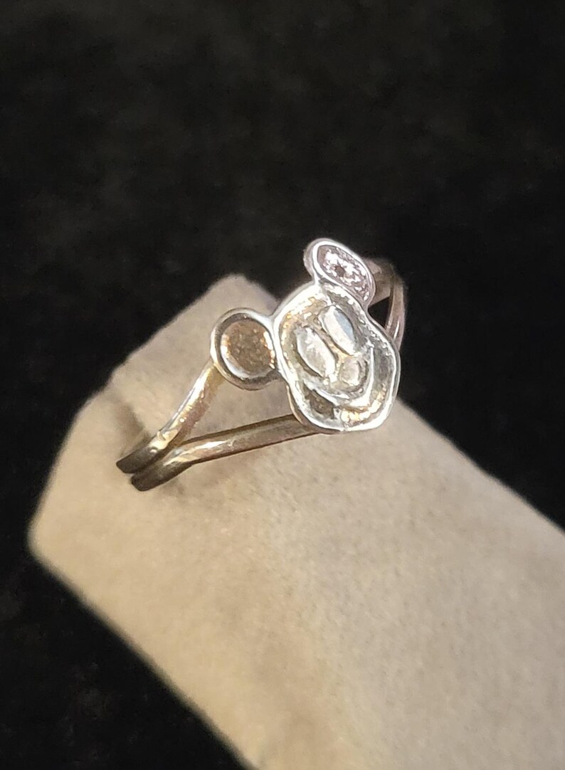 Puede incluir: Un anillo de metal plateado que presenta una cabeza de personaje estilizada en relieve con dos orejas redondeadas. La banda del anillo es delgada y se divide en dos secciones al conectar con el dise&ntilde;o central, creando un aspecto simple y abierto.