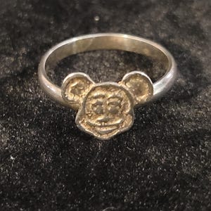 Pu&ograve; includere: Un anello in metallo color argento con un ciondolo centrale a forma di testa di topo dei cartoni animati, con orecchie rotonde, un ampio sorriso e dettagli strutturati sul viso. La fascia &egrave; liscia, circolare e lucida, unita alla base della testa.