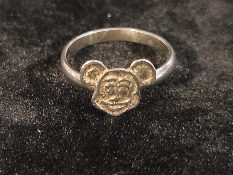 Pu&ograve; includere: Un anello in metallo color argento con un ciondolo centrale a forma di testa di topo dei cartoni animati, con orecchie rotonde, un ampio sorriso e dettagli strutturati sul viso. La fascia &egrave; liscia, circolare e lucida, unita alla base della testa.