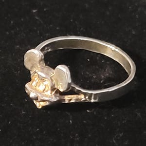 Puede incluir: Anillo de plata con dise&ntilde;o de Mickey Mouse. El anillo tiene una banda plateada y una cabeza de Mickey Mouse dorada. El anillo est&aacute; sobre un fondo oscuro.