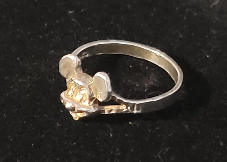 Puede incluir: Anillo de plata con dise&ntilde;o de Mickey Mouse. El anillo tiene una banda plateada y una cabeza de Mickey Mouse dorada. El anillo est&aacute; sobre un fondo oscuro.