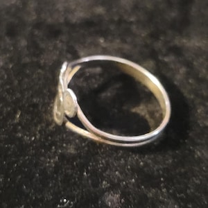Puede incluir: Un anillo de metal plateado con un dise&ntilde;o abierto de dos extremos. Un extremo de la banda termina en un peque&ntilde;o disco circular plano, mientras que el otro extremo presenta una forma met&aacute;lica ligeramente m&aacute;s grande, alargada y redondeada.