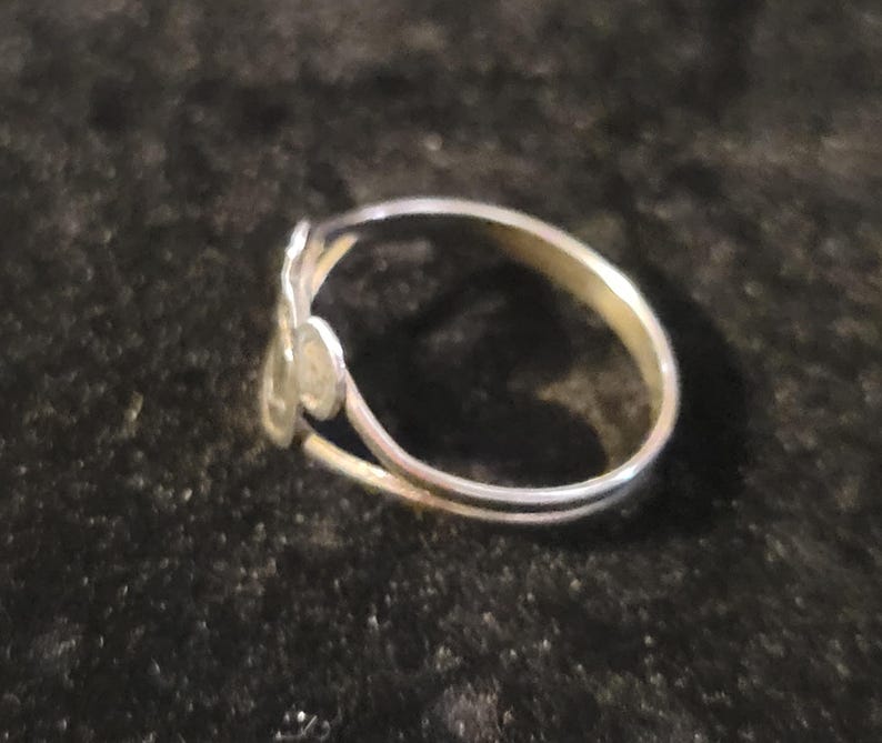 Puede incluir: Un anillo de metal plateado con un dise&ntilde;o abierto de dos extremos. Un extremo de la banda termina en un peque&ntilde;o disco circular plano, mientras que el otro extremo presenta una forma met&aacute;lica ligeramente m&aacute;s grande, alargada y redondeada.