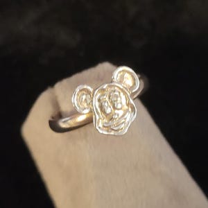 Pu&ograve; includere: Un anello in metallo color argento caratterizzato da un design in rilievo a forma di testa di personaggio stilizzata, con due orecchie arrotondate e un volto centrale. Il gambo dell'anello &egrave; liscio e circolare, con una finitura metallica.
