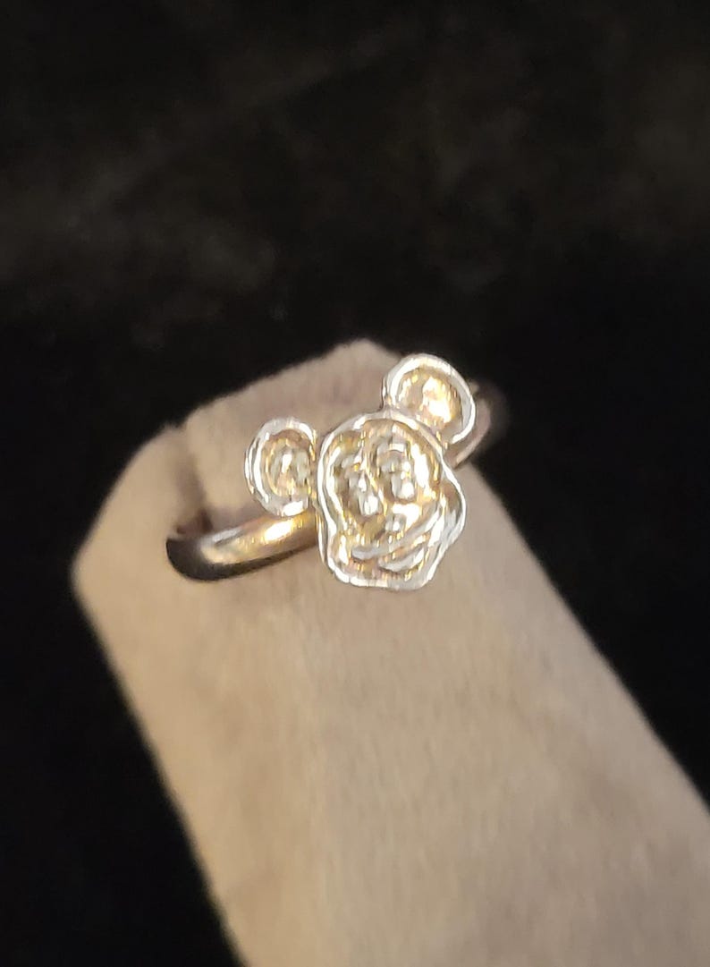 Pu&ograve; includere: Un anello in metallo color argento caratterizzato da un design in rilievo a forma di testa di personaggio stilizzata, con due orecchie arrotondate e un volto centrale. Il gambo dell'anello &egrave; liscio e circolare, con una finitura metallica.