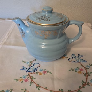 May include: A light blue ceramic teapot featuring horizontal ribbed detailing, a matching lid, and gold-colored floral accents on the body and lid. The teapot has a curved handle and a spout, with gold trim along the lid edge and spout rim.