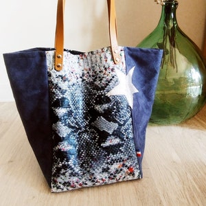 Peut inclure: Un sac cabas en toile bleu avec un motif peau de serpent et une étoile argentée. Le sac a des anses en cuir marron et un petit drapeau français.