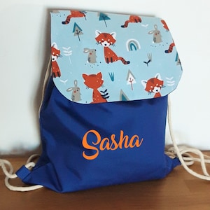 May include: Blue drawstring backpack with a light blue flap featuring a red panda and bunny pattern. The backpack has the name "Sasha" embroidered on the front in orange.