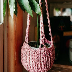 May include: A pink crocheted hanging planter with a green plant inside. The planter is made with a delicate stitch pattern and has a white crocheted strap.