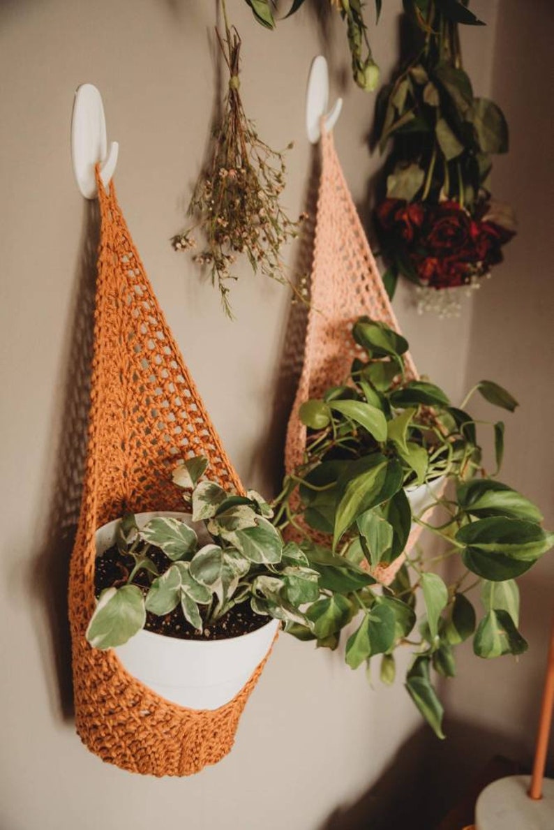 May include: Two handmade crochet plant hangers in shades of orange and peach, each holding a white pot with greenery. The hangers are suspended from white wall hooks. Dried flowers and roses hang in the background.