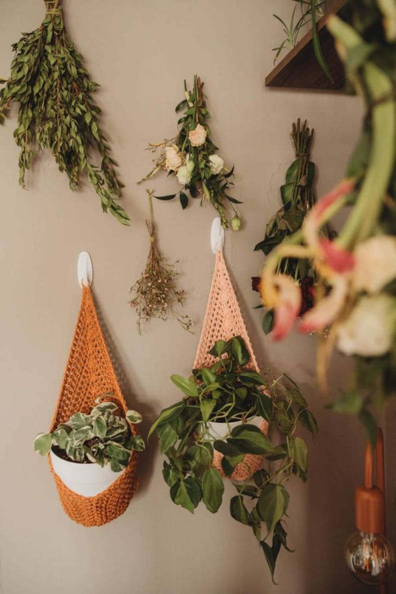 May include: A collection of hanging planters and dried flowers against a neutral wall. Two crochet plant holders in orange and peach colors hold potted plants. Various dried flowers and greenery are also displayed.