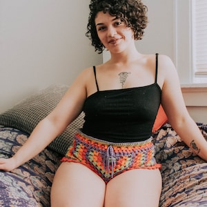 May include: A woman with curly brown hair is wearing a black tank top and colorful crochet shorts. She is sitting on a patterned bed with a pillow behind her.