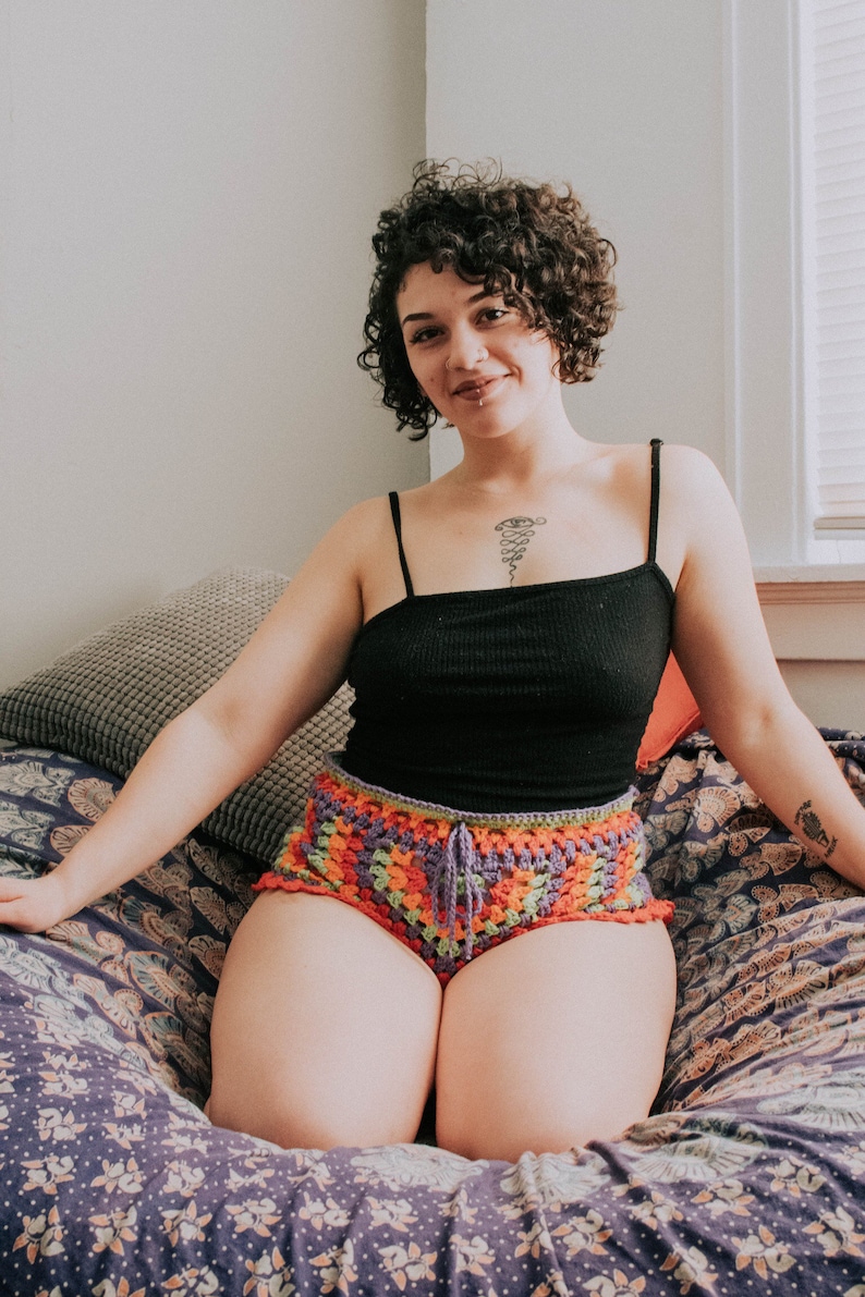 May include: A woman with curly brown hair is wearing a black tank top and colorful crochet shorts. She is sitting on a patterned bed with a pillow behind her.