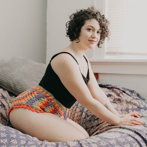 May include: A woman wearing a black tank top and a colorful crocheted bikini bottom sits on a floral patterned bed.