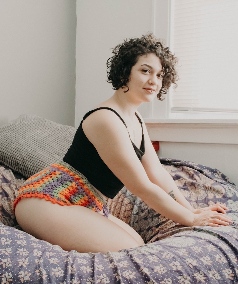 May include: A woman wearing a black tank top and a colorful crocheted bikini bottom sits on a floral patterned bed.