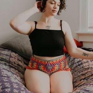 May include: A person wearing a black tank top and colorful crochet shorts sits on a patterned cushion. They have short curly hair and a tattoo on their chest.