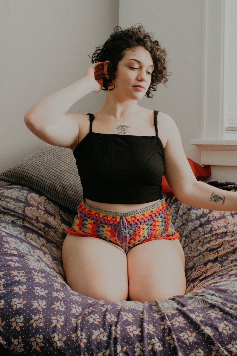 May include: A person wearing a black tank top and colorful crochet shorts sits on a patterned cushion. They have short curly hair and a tattoo on their chest.