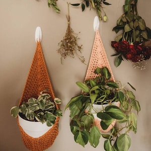 May include: Two handmade crochet plant hangers in orange and pink, each holding a white pot with greenery. Dried flowers and roses hang on the wall above the plant hangers, creating a natural, bohemian aesthetic.