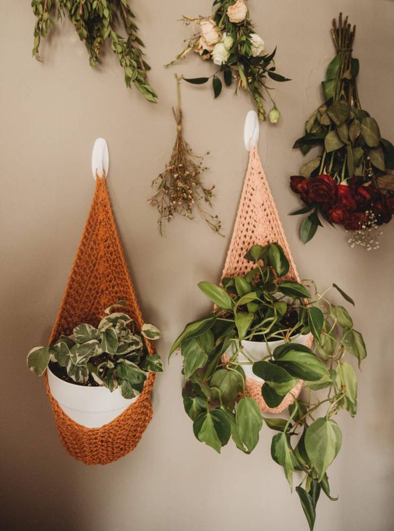 May include: Two handmade crochet plant hangers in orange and pink, each holding a white pot with greenery. Dried flowers and roses hang on the wall above the plant hangers, creating a natural, bohemian aesthetic.