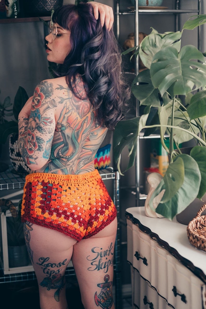 May include: A woman with purple hair and tattoos is wearing a crocheted orange and red bikini bottom. The bikini bottom has a square pattern. The woman is standing in front of a plant and a dresser. The text "Loose Lips Sink Ships" is tattooed on her leg.