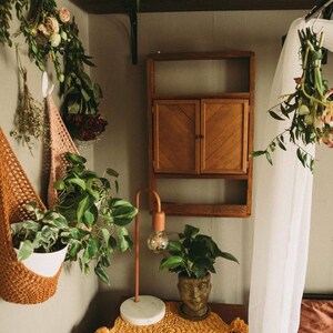 May include: A cozy interior scene featuring a wooden cabinet, a copper lamp, and a small table adorned with a yellow crocheted runner. Potted plants and hanging dried flowers add a natural touch to the space.