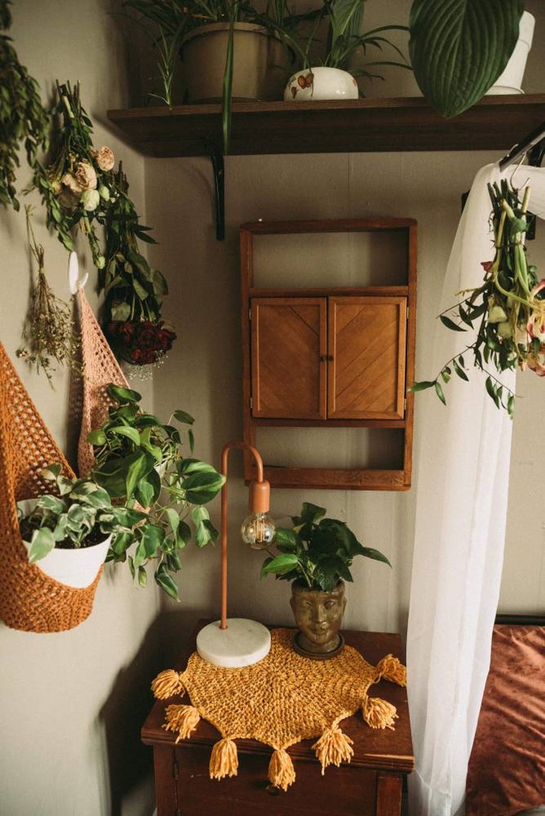 May include: A cozy interior scene featuring a wooden cabinet, a copper lamp, and a small table adorned with a yellow crocheted runner. Potted plants and hanging dried flowers add a natural touch to the space.