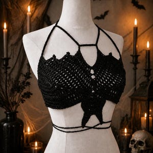 May include: Black crochet halter top with a bat-wing design. The top features a halter neck and a tie closure around the waist. The garment is displayed on a mannequin, with a Halloween-themed background including candles and bat silhouettes.