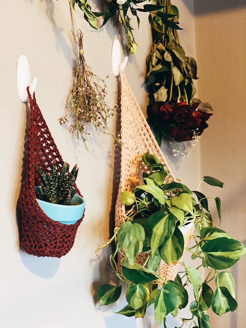 May include: Two handmade crochet plant hangers in shades of burgundy and peach, each holding a potted plant. The burgundy hanger contains a succulent in a blue pot, while the peach one displays a trailing green plant. Dried flowers hang nearby.