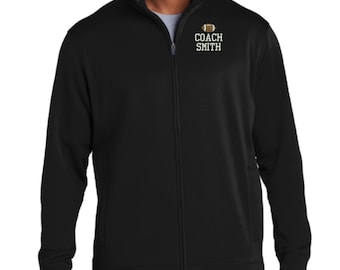 May include: A black full-zip jacket with a mock neck collar. On the upper left chest, there is a graphic of a brown football positioned above the white text that reads COACH SMITH. The jacket features long sleeves and a central vertical zipper.