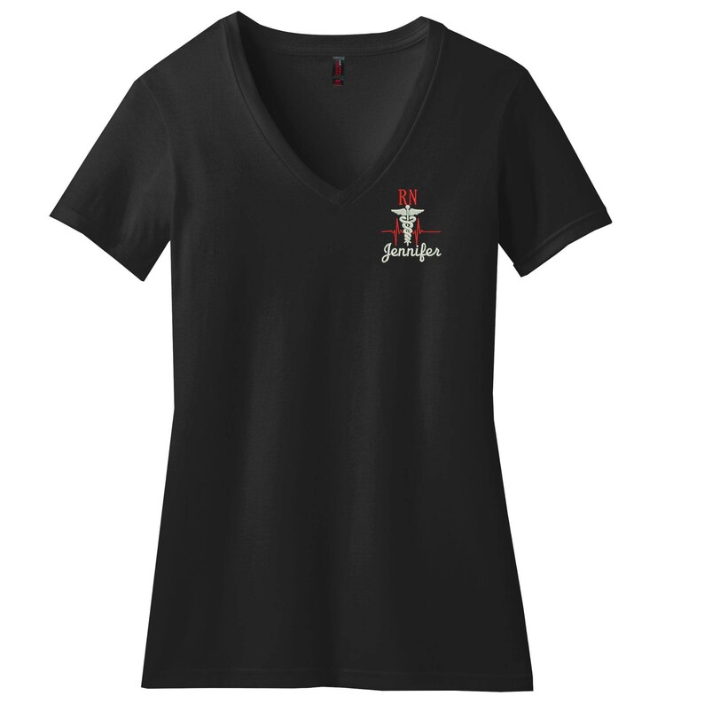 May include: A black V-neck t-shirt featuring an embroidered design on the left chest. The design includes a red heartbeat line, a white caduceus symbol, the red letters RN, and the name Jennifer in white script lettering below the symbol.