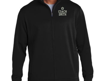 May include: A black full-zip jacket with a mock neck collar. On the upper left chest, there is a white graphic of a soccer ball above the text "COACH SMITH" in a bold, block-style font. The jacket features long sleeves and side pockets.