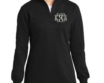 May include: A black long-sleeve pullover with a quarter-zip closure at the collar. The garment features a white embroidered monogram on the upper left chest area, consisting of three stylized, intertwined letters in a decorative script font.
