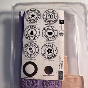 May include: A clear plastic container holds a stamp set. The set includes circular stamps with phrases like "Wishing You The Best" and "Happy Holidays." Purple rubber stamps and wooden blocks are also visible.