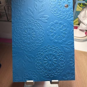 May include: A rectangular blue cardstock sheet featuring a raised, embossed floral pattern with intricate circular blooms and leaf motifs. The cardstock has a matte texture and includes a single circular hole punched near the top right corner.