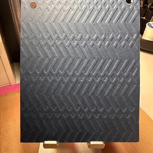 May include: A rectangular dark blue cardstock sheet featuring a debossed herringbone and diamond pattern. The sheet has two circular holes punched near the top corners and is displayed on a silver metal stand, showcasing a textured surface.