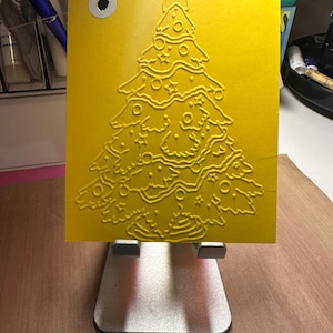 May include: A bright yellow rectangular cardstock featuring a raised, embossed design of a Christmas tree decorated with stars and circular ornaments. The card has a single white-reinforced hole punch in the top left corner and is displayed on a silver stand.