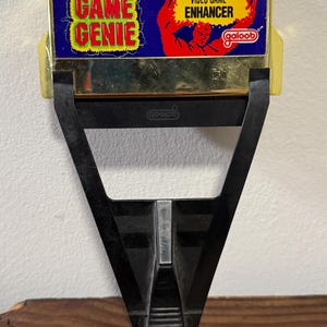 Puede incluir: Un potenciador de videojuegos Game Genie vintage. El dispositivo tiene un marco de plástico negro con una sección superior dorada. La sección superior presenta el texto "GAME GENIE" y "VIDEO GAME ENHANCER". También es visible la marca "galoob".