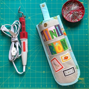 Puede incluir: Una bolsa de tela azul claro con la palabra "mini plancha" impresa en ella en letras de colores. La bolsa tiene varios bolsillos y está llena de artículos de costura, incluyendo tijeras y un pequeño cojín de alfileres rojo.