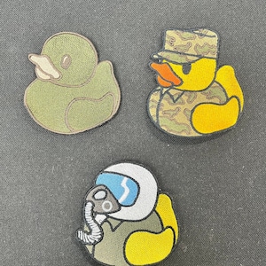 May include: Three embroidered iron-on patches featuring yellow rubber duckies in different outfits. The first patch shows a green duck wearing a camouflage hat. The second patch shows a yellow duck wearing a camouflage uniform. The third patch shows a yellow duck wearing a white helmet and a green flight suit.