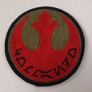 May include: A red embroidered Rebel Alliance symbol patch with a black border and green background. The text "REBEL ALLIANCE" is embroidered around the symbol in black.