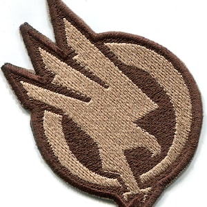May include: A brown and tan embroidered patch with a stylized eagle design. The eagle is facing left and has its wings spread. The patch is circular and has a brown border.