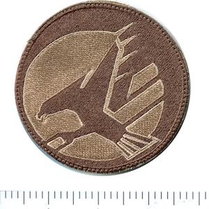 May include: A brown circular patch with a tan embroidered eagle design. The eagle is facing right and has its wings spread. The patch has a brown stitched border.