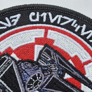 May include: Black and red embroidered patch with a TIE fighter silhouette and the text "TIE/IN" in white.