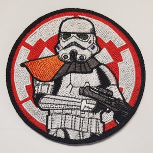 Może przedstawiać: Naszywka z haftem przedstawiająca białego szturmowca z Gwiezdnych Wojen na tle czerwono-białego symbolu Imperium.