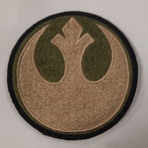 May include: Embroidered patch featuring the Rebel Alliance symbol from Star Wars. The patch is circular, with a tan emblem on a green background, outlined in black. The design is detailed and suitable for application on clothing or accessories.