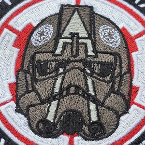 May include: A close-up of a gray and black embroidered patch featuring a stylized helmet with a red and white circular border. The helmet has a black visor and a gray and white design.