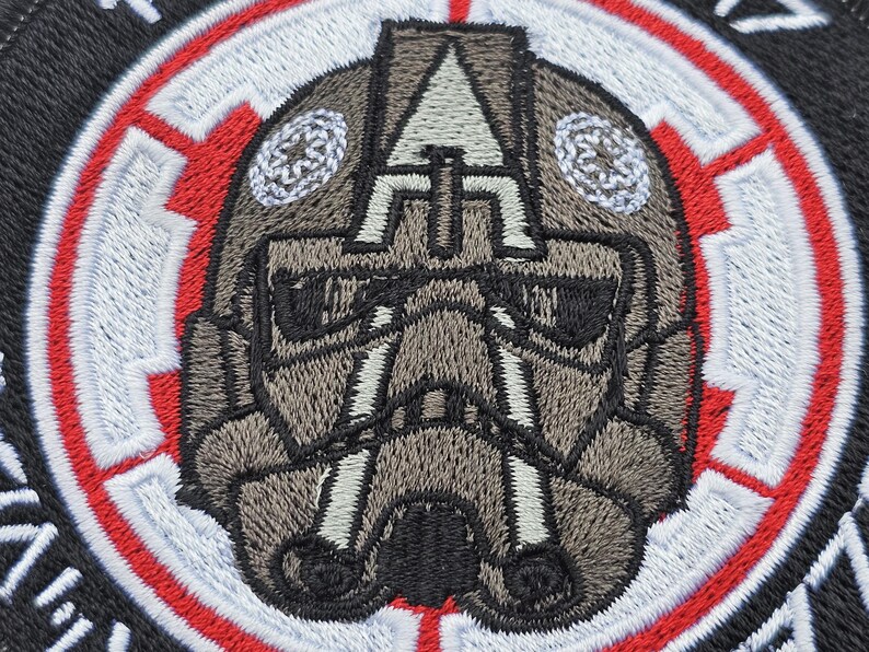 May include: A close-up of a gray and black embroidered patch featuring a stylized helmet with a red and white circular border. The helmet has a black visor and a gray and white design.